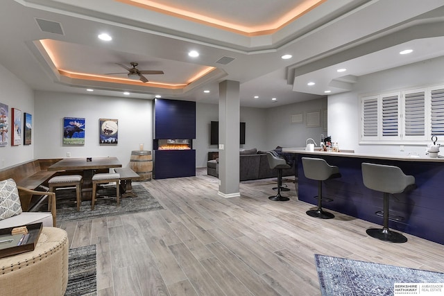 interior space with ceiling fan, a tray ceiling, a fireplace, bar area, and light wood-type flooring