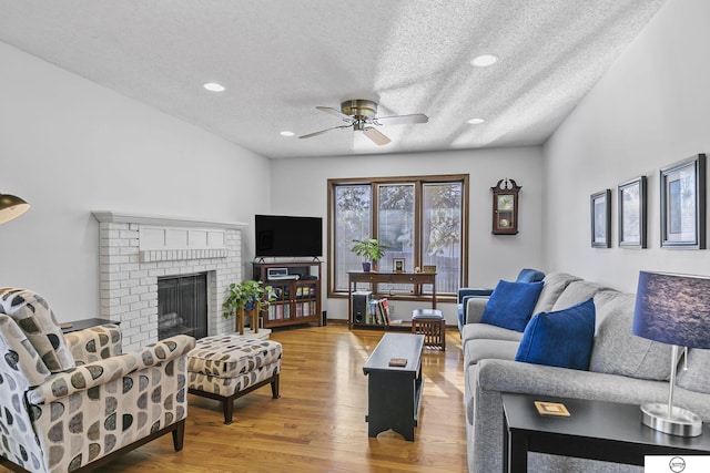 living room with a brick fireplace, wood-type flooring, a textured ceiling, and ceiling fan