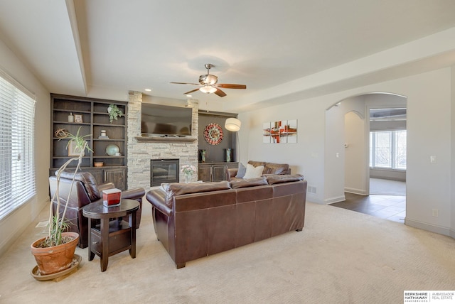 living room featuring ceiling fan, baseboards, carpet, a stone fireplace, and arched walkways