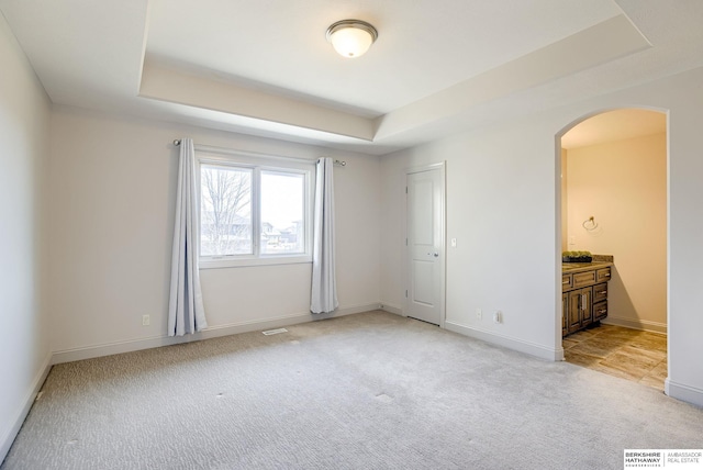 unfurnished bedroom with a raised ceiling, light colored carpet, arched walkways, and baseboards