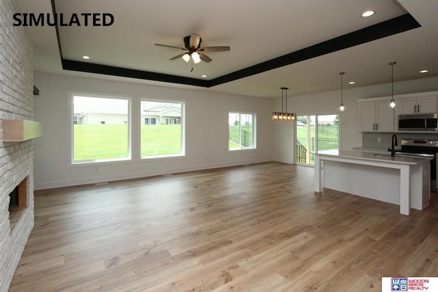 unfurnished living room featuring a fireplace, a raised ceiling, ceiling fan with notable chandelier, and light wood-type flooring