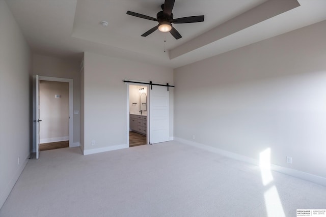 unfurnished bedroom with ensuite bath, ceiling fan, a tray ceiling, light carpet, and a barn door