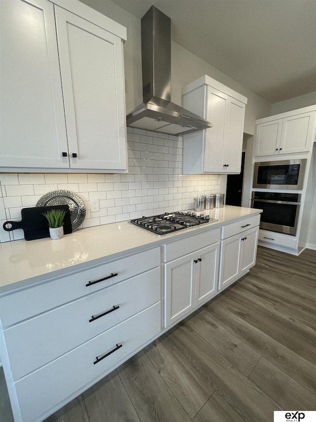 kitchen with appliances with stainless steel finishes, wood-type flooring, white cabinets, decorative backsplash, and wall chimney exhaust hood