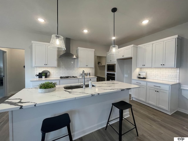 kitchen featuring white cabinets, a center island with sink, stainless steel microwave, and wall chimney exhaust hood