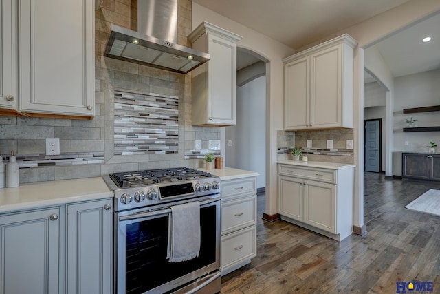 kitchen featuring dark wood-style floors, arched walkways, light countertops, stainless steel range with gas stovetop, and wall chimney exhaust hood