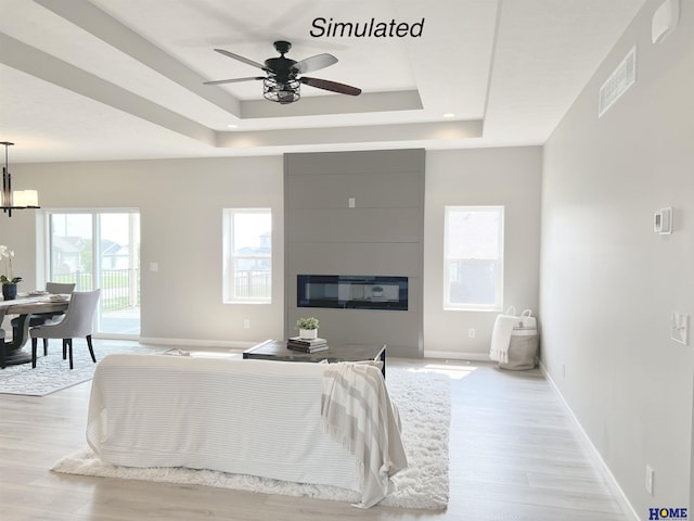 interior space with a raised ceiling, a large fireplace, ceiling fan with notable chandelier, and light wood-type flooring