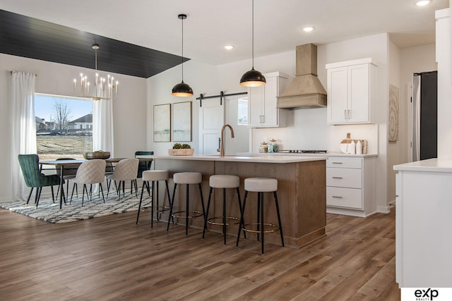 kitchen with custom exhaust hood, white cabinetry, decorative light fixtures, a center island with sink, and a barn door