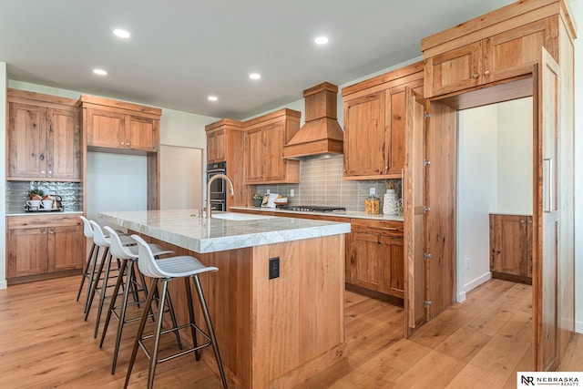 kitchen with sink, custom exhaust hood, a kitchen breakfast bar, a kitchen island with sink, and light hardwood / wood-style floors