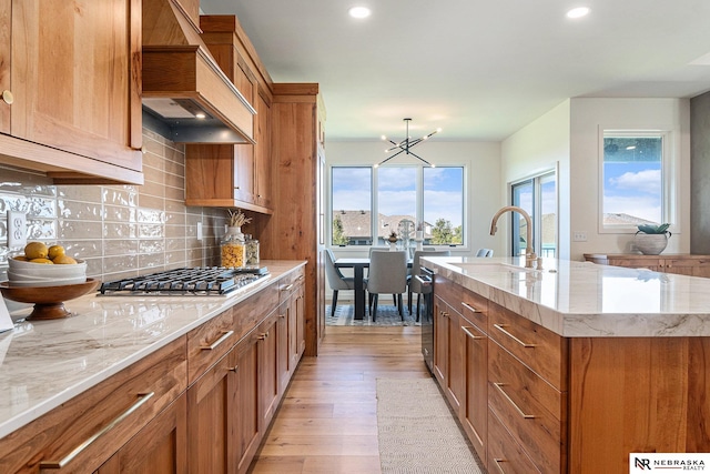 kitchen featuring stainless steel gas cooktop, a kitchen island with sink, custom range hood, light hardwood / wood-style floors, and decorative backsplash