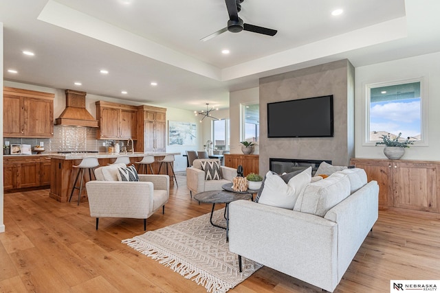 living room with a raised ceiling, ceiling fan with notable chandelier, a fireplace, and light hardwood / wood-style flooring