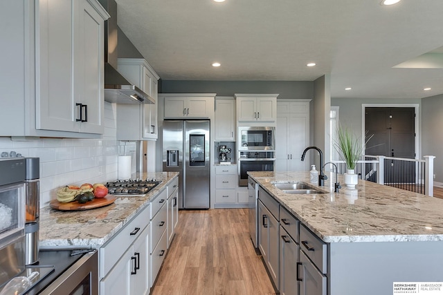 kitchen with appliances with stainless steel finishes, an island with sink, white cabinets, light wood-type flooring, and wall chimney exhaust hood