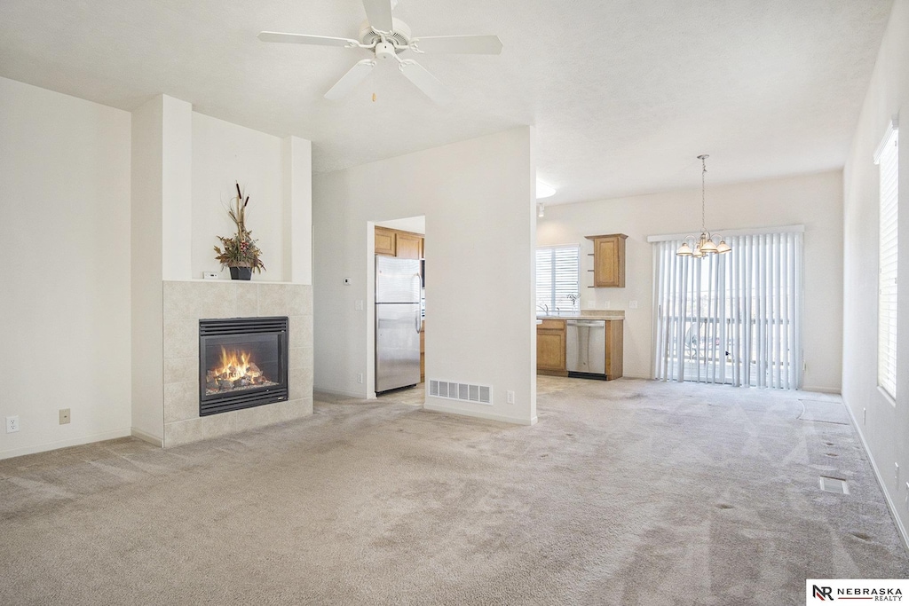 unfurnished living room with a tile fireplace, ceiling fan with notable chandelier, and light colored carpet