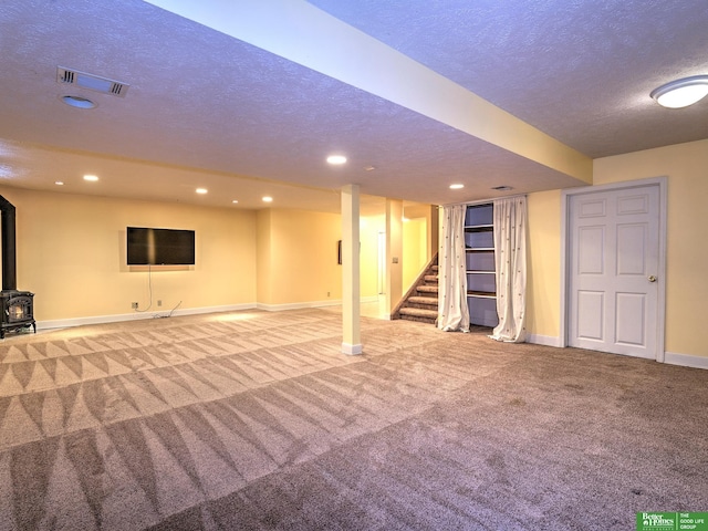 finished below grade area featuring visible vents, a wood stove, stairs, a textured ceiling, and carpet floors