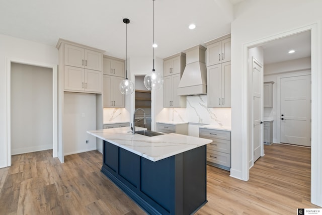 kitchen featuring sink, premium range hood, a kitchen island with sink, light stone countertops, and light wood-type flooring