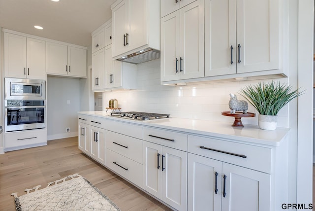 kitchen featuring appliances with stainless steel finishes, ventilation hood, white cabinetry, decorative backsplash, and light wood-type flooring