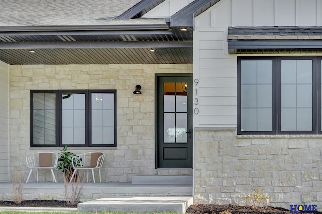 entrance to property with board and batten siding, stone siding, roof with shingles, and covered porch