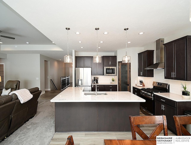 kitchen with open floor plan, a kitchen island with sink, a sink, built in appliances, and wall chimney exhaust hood