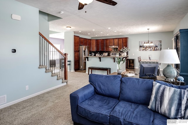 living room featuring stairway, light carpet, a textured ceiling, baseboards, and ceiling fan with notable chandelier