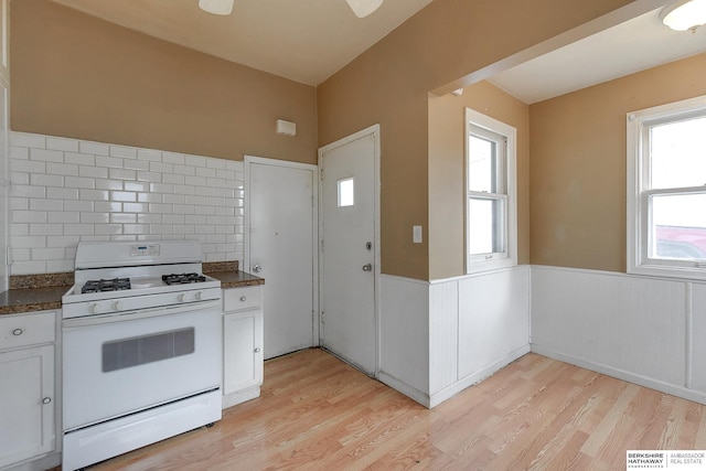 kitchen with white range with gas stovetop, decorative backsplash, a wainscoted wall, light wood-style floors, and white cabinetry