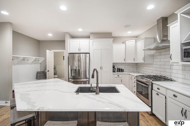 kitchen with a center island with sink, wall chimney exhaust hood, light stone counters, stainless steel appliances, and a sink