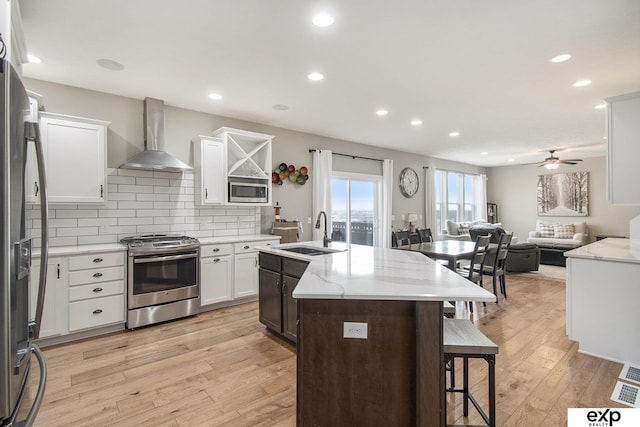 kitchen with open floor plan, stainless steel appliances, wall chimney range hood, white cabinetry, and a sink