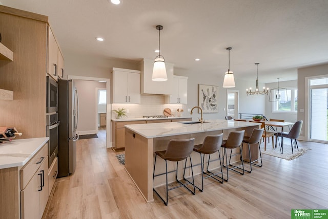 kitchen with light stone counters, a kitchen island with sink, a sink, white cabinets, and hanging light fixtures