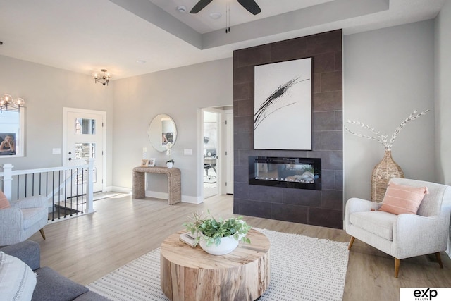 living room with a tile fireplace, ceiling fan with notable chandelier, baseboards, light wood-style floors, and a tray ceiling