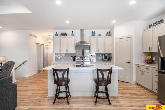 kitchen featuring light countertops, a barn door, an island with sink, and wall chimney range hood