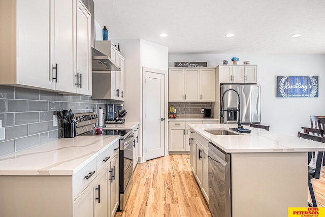 kitchen with appliances with stainless steel finishes, a kitchen island with sink, light wood-style floors, and wall chimney range hood