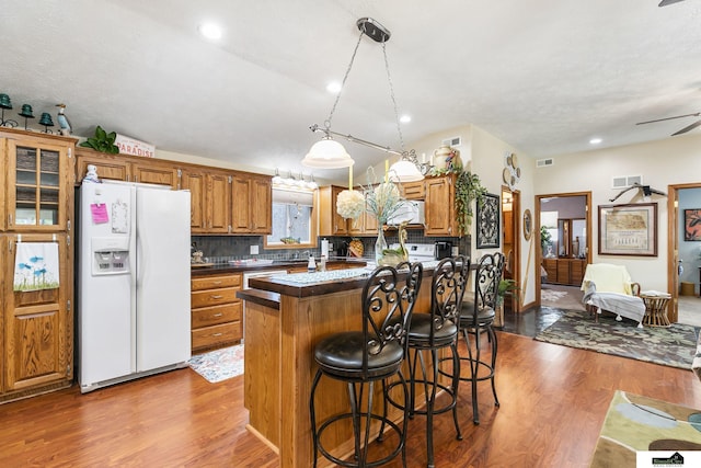 kitchen with brown cabinetry, dark countertops, a breakfast bar area, white fridge with ice dispenser, and pendant lighting