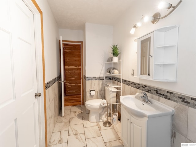 half bathroom featuring wainscoting, toilet, marble finish floor, vanity, and tile walls