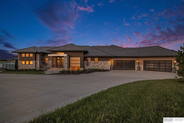 prairie-style house featuring a garage, stone siding, driveway, and a yard