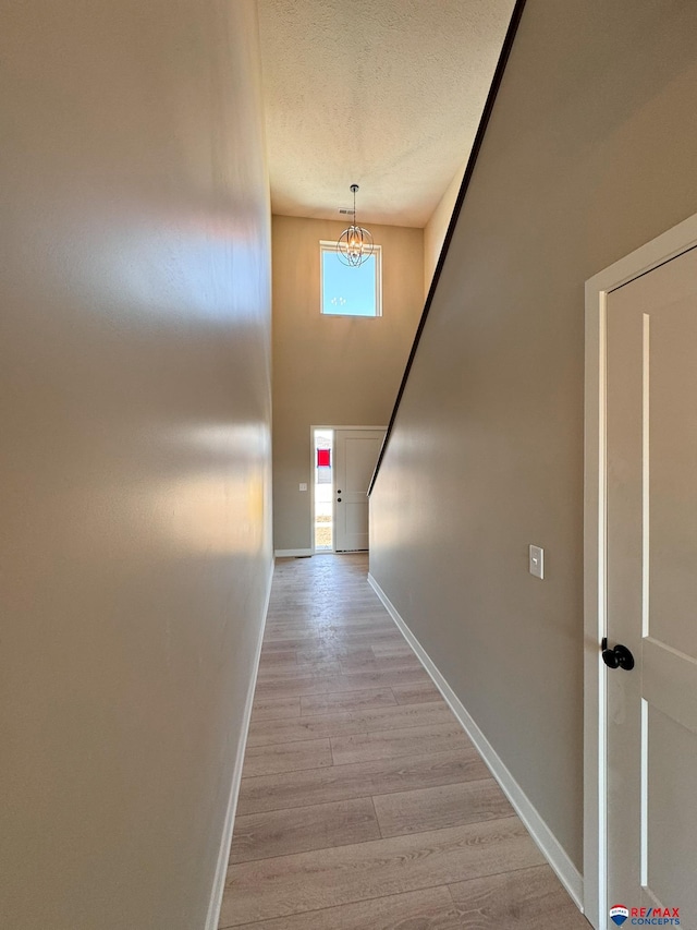 hall with a high ceiling, an inviting chandelier, a textured ceiling, light wood-type flooring, and baseboards