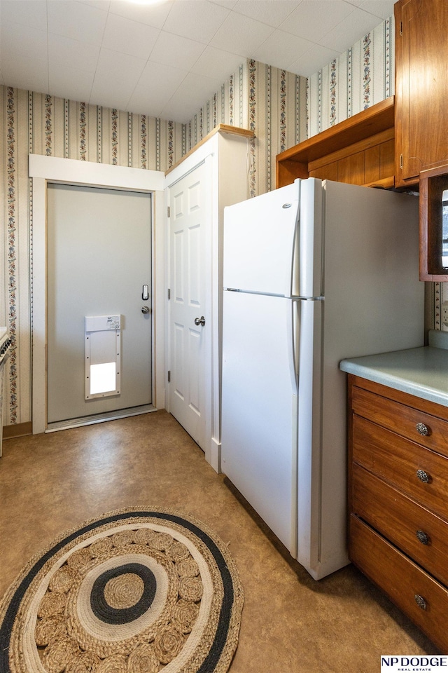 kitchen with brown cabinetry, freestanding refrigerator, light countertops, and wallpapered walls