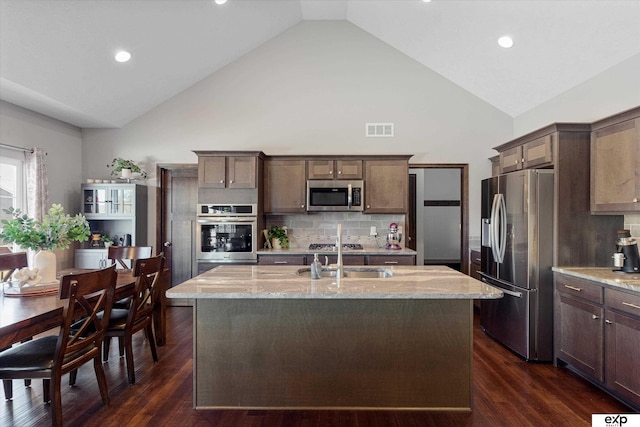 kitchen with light stone counters, a center island with sink, stainless steel appliances, visible vents, and a sink