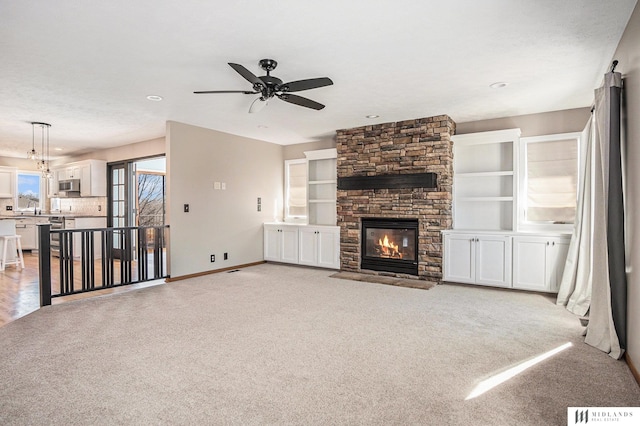 unfurnished living room with light colored carpet, ceiling fan, a stone fireplace, and baseboards