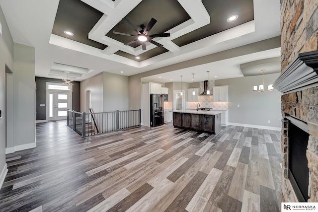 unfurnished living room featuring a stone fireplace, ceiling fan with notable chandelier, coffered ceiling, wood finished floors, and baseboards