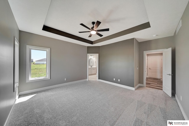 unfurnished bedroom with ceiling fan, ensuite bathroom, light carpet, baseboards, and a raised ceiling