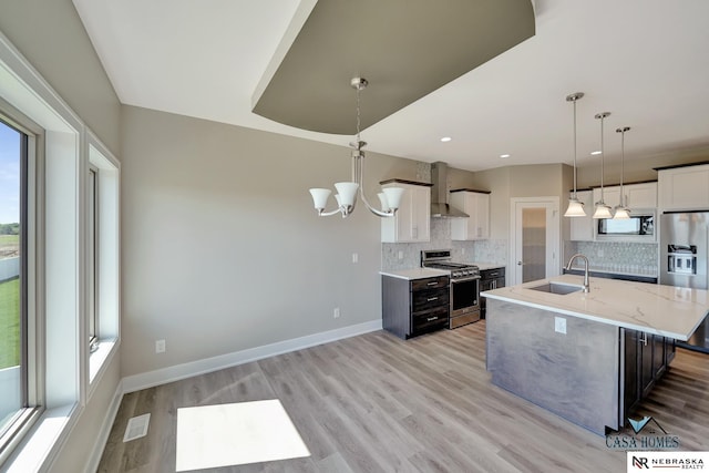 kitchen featuring an island with sink, stainless steel appliances, wall chimney range hood, white cabinetry, and pendant lighting