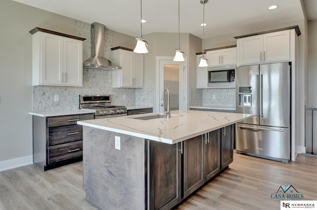 kitchen with pendant lighting, high end appliances, a sink, an island with sink, and wall chimney exhaust hood