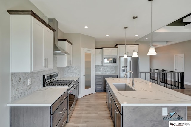 kitchen featuring a sink, hanging light fixtures, wall chimney range hood, appliances with stainless steel finishes, and an island with sink