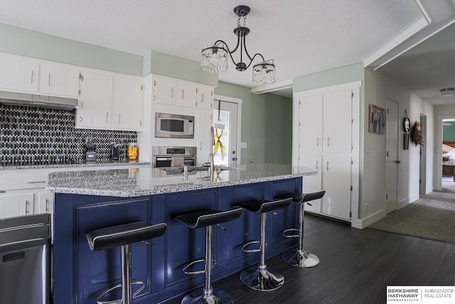 kitchen with pendant lighting, dark wood finished floors, a center island with sink, stainless steel appliances, and white cabinetry