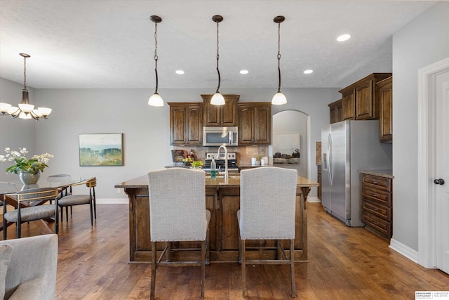 kitchen with arched walkways, a kitchen island with sink, appliances with stainless steel finishes, backsplash, and dark wood-style floors