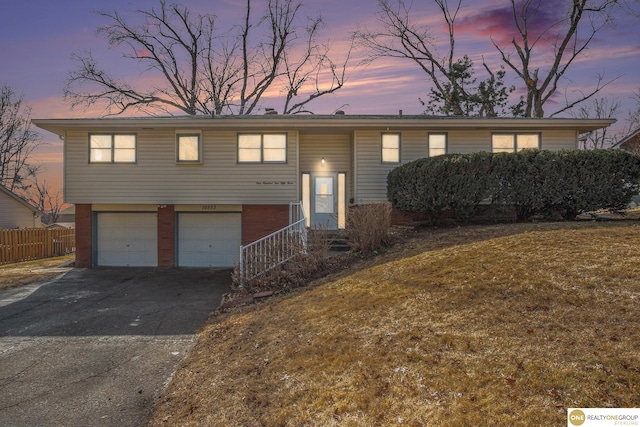 bi-level home featuring aphalt driveway, a garage, brick siding, fence, and a lawn