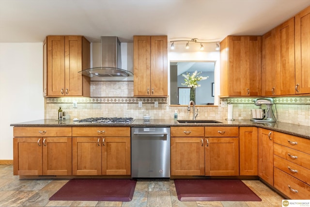 kitchen with brown cabinets, dark stone countertops, stainless steel appliances, wall chimney range hood, and a sink