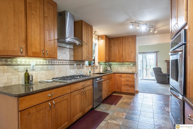 kitchen with tasteful backsplash, brown cabinetry, wall chimney exhaust hood, appliances with stainless steel finishes, and a sink