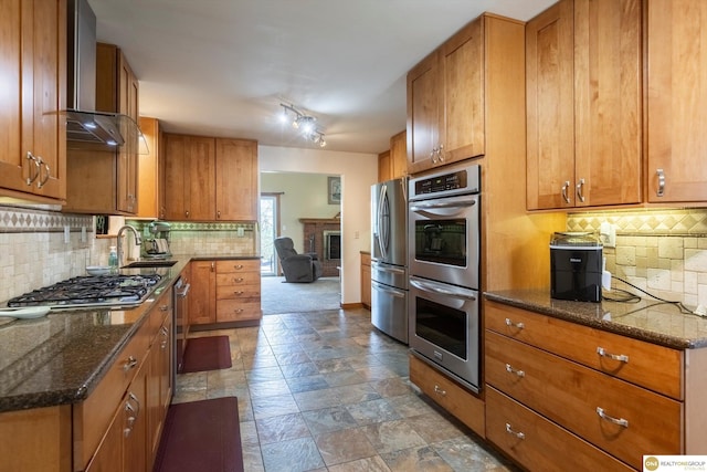 kitchen with brown cabinets, wall chimney range hood, stainless steel appliances, and backsplash