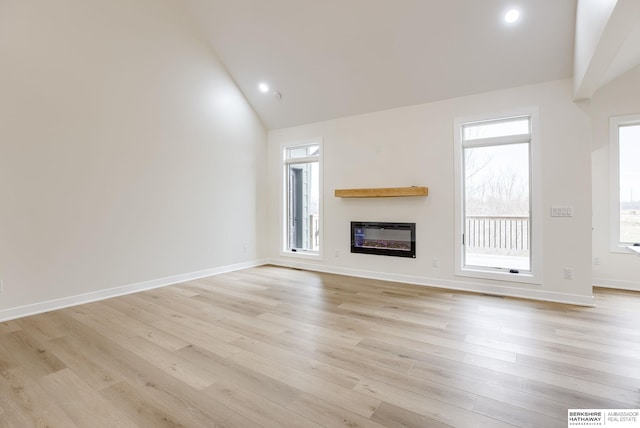 unfurnished living room with high vaulted ceiling, light wood-type flooring, a glass covered fireplace, and baseboards