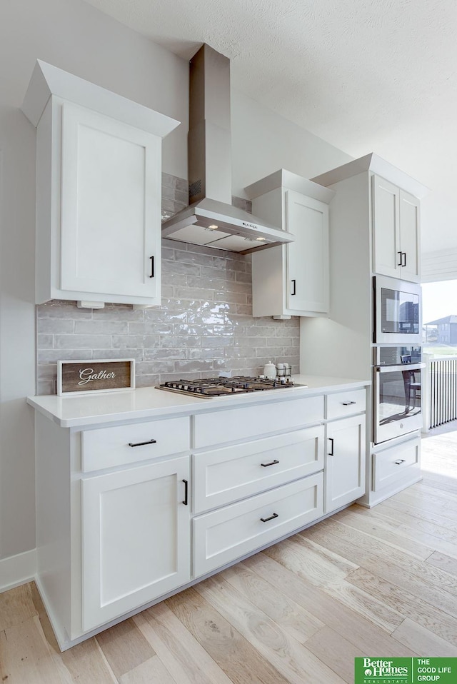 kitchen with white cabinetry, light countertops, appliances with stainless steel finishes, light wood-type flooring, and wall chimney exhaust hood