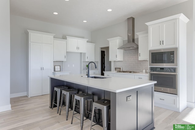 kitchen with stainless steel appliances, a sink, wall chimney exhaust hood, light wood finished floors, and a large island with sink